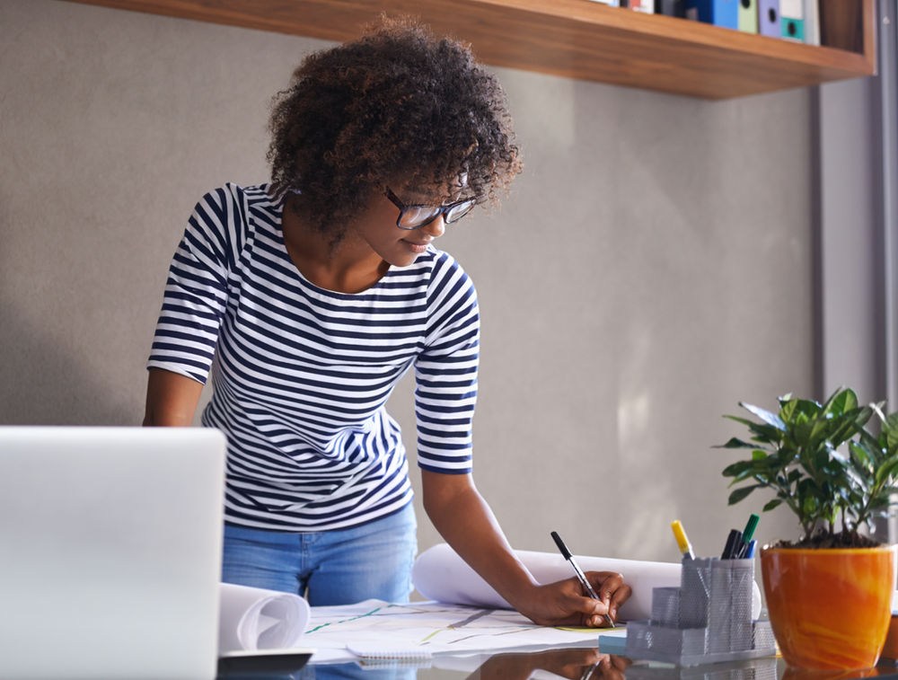 Woman in a striped shirt writes on papers at a desk, making a financial plan, with a laptop and a plant in an orange pot. Shelves with books in the background.