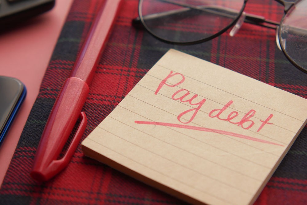 Note with "Pay debt" on a lined sticky note, red pen, and glasses on a red plaid fabric background. Mood suggests urgency.