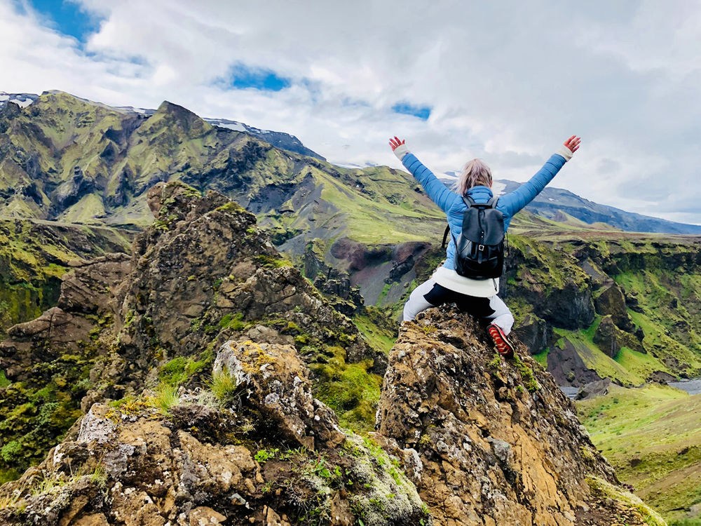 Person in a blue jacket celebrates atop a rocky hill, arms raised, celebrating with lush green mountains in the background under a cloudy sky.