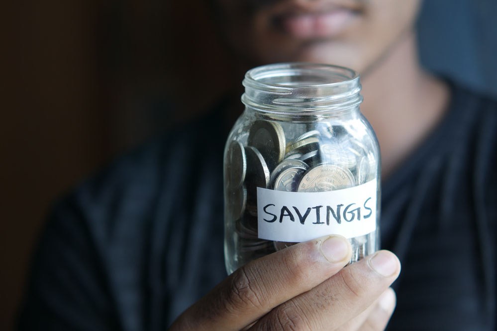 Person holding a jar labeled "SAVINGS" filled with coins. Focus on hand and jar, with blurred background creating a thoughtful mood.