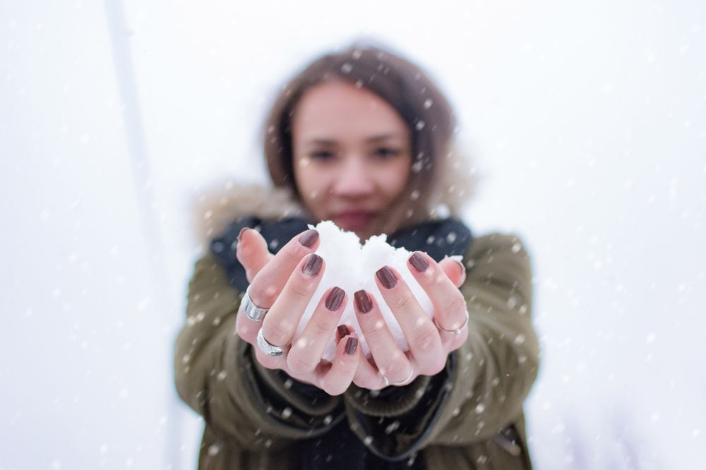 Woman in winter coat holding snowball with both hands. Snow falls gently around, with nails painted dark red. Blurry snowy background.