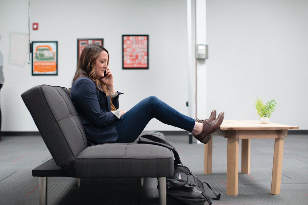Woman sitting on gray sofa, talking on phone with feet on a table. Bright room with framed posters and a small plant on the table.