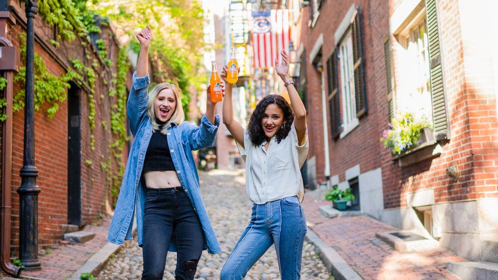Two women cheerfully raise bottles on a sunny cobblestone street with brick buildings and an American flag in the background.