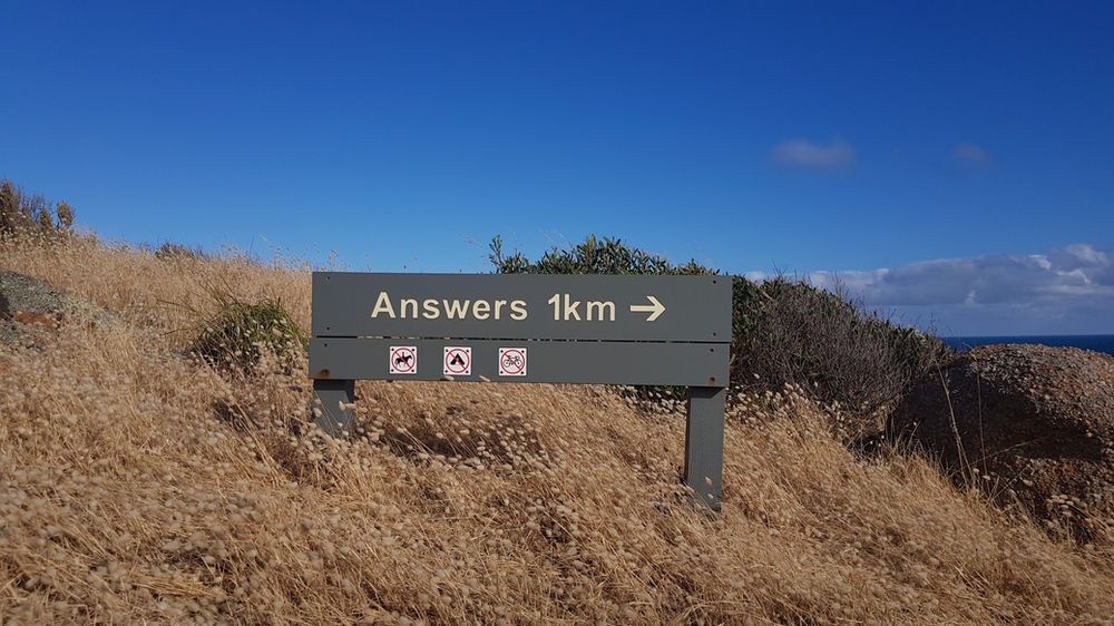 Sign on a grassy hill reads "Answers 1km" with a right arrow. Clear blue sky and distant ocean visible.
